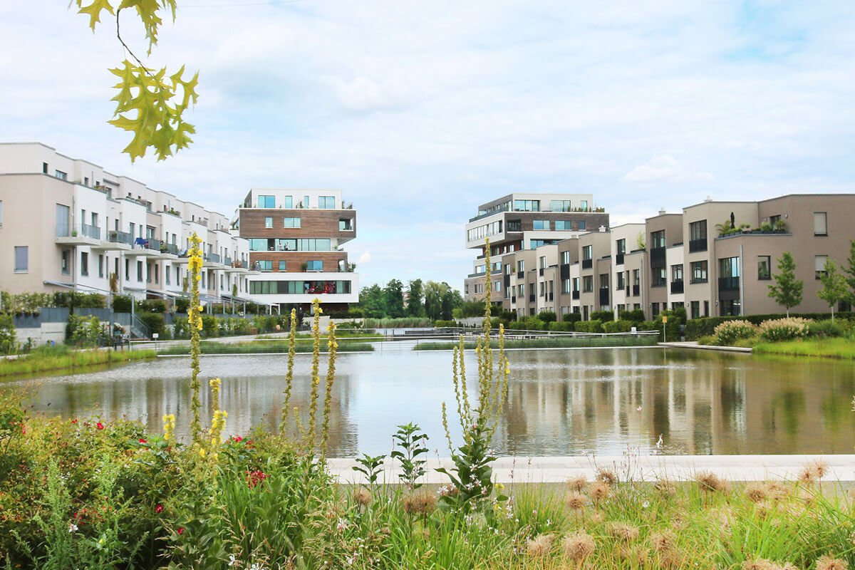Ein mehrstöckige moderne Wohngebäude mit hellbeiger Fassade und mehreren verglasten Balkonen, das von einer gepflegten Grünanlage mit Bäumen umgeben ist. Im Vordergrund sieht man einen Teich. Der klare blaue Himmel und das Sonnenlicht deuten auf einen schönen, sonnigen Tag hin.