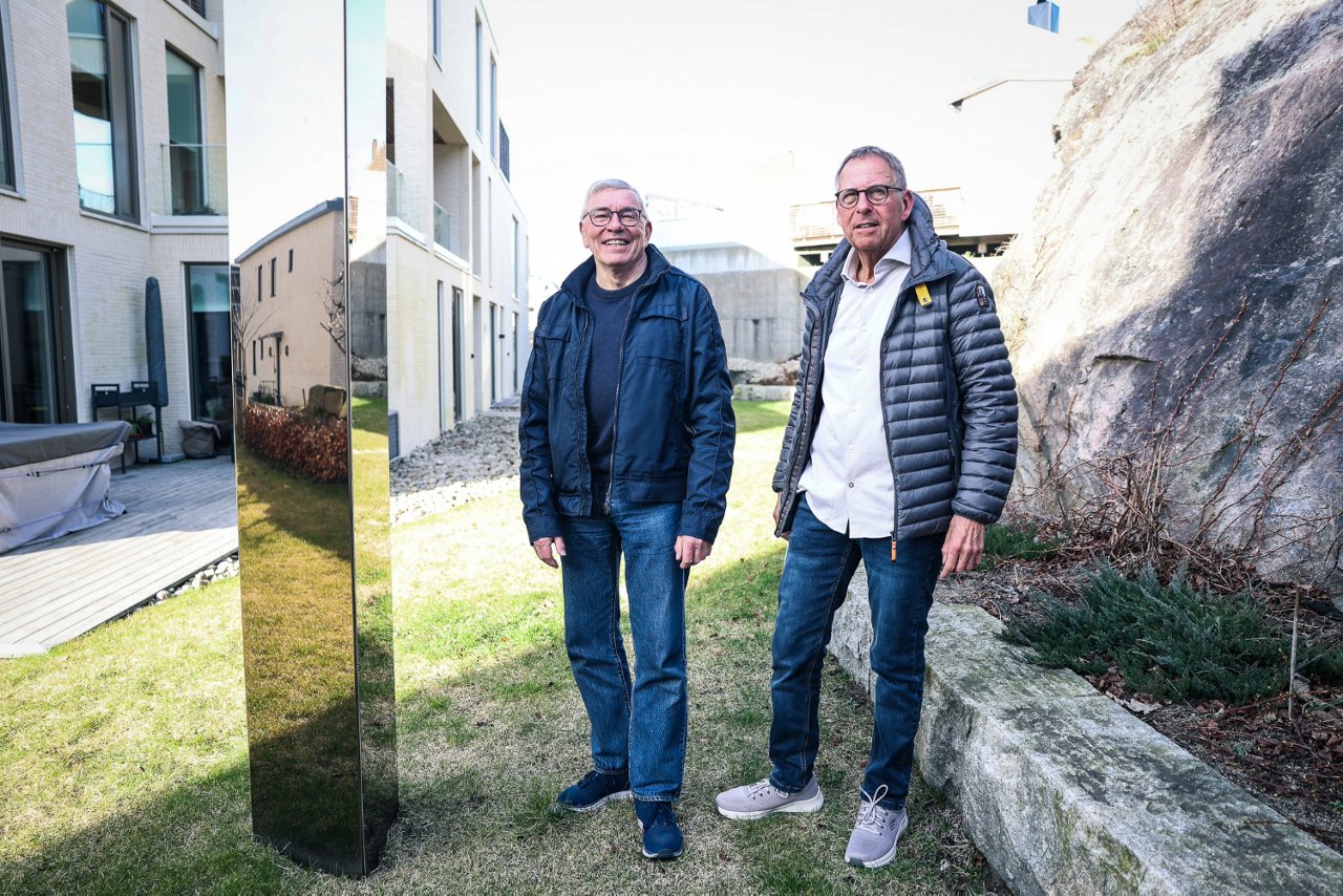 To menn står utenfor en bygning, grønt gress og solen skinner.