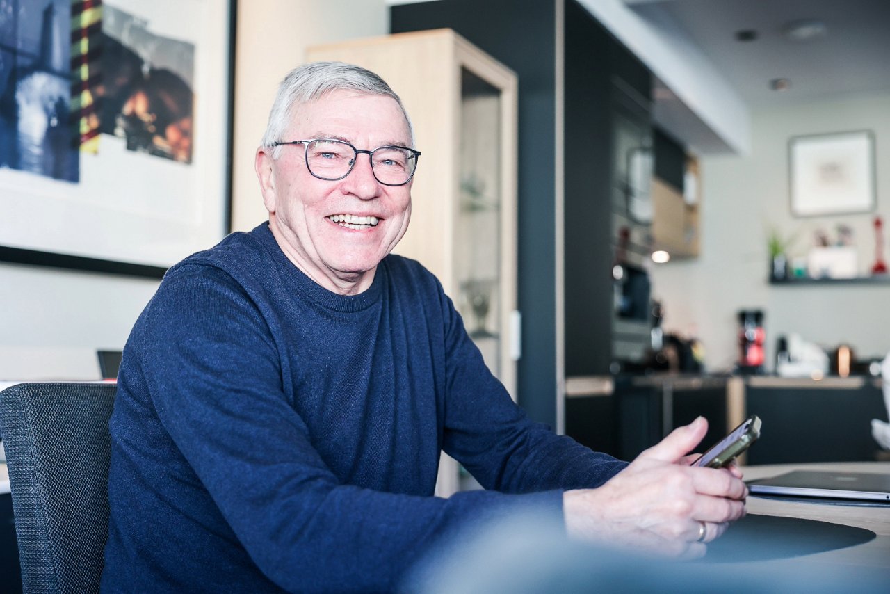 En mann med briller som smiler mens han holder en mobiltelefon i hånden.