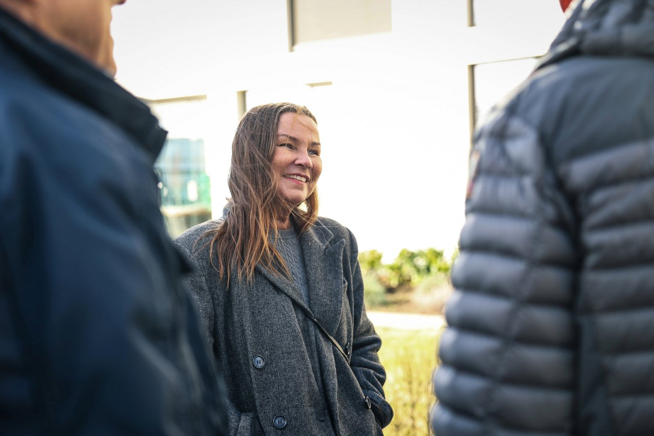 En dame som smiler og har hendene i lommene. Hun ser ut til å være i en samtale med to personer, hvor vi ser ryggen deres.