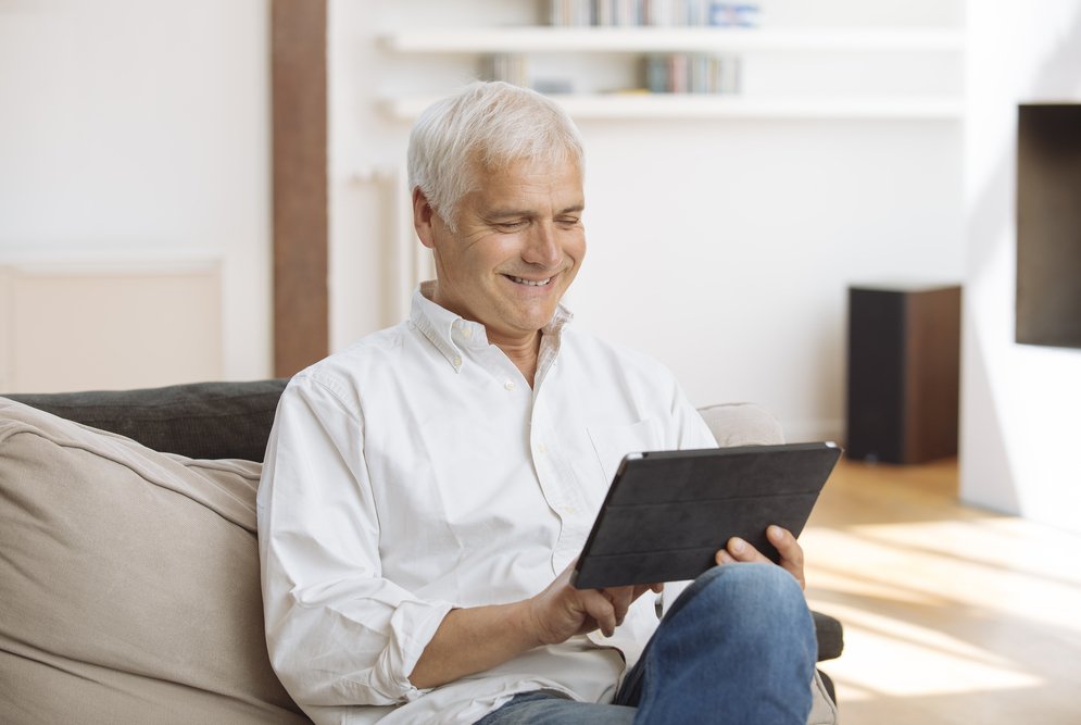 Hombre de mediana edad y pelo blanco sentado en un sofá, sosteniendo y mirando una tablet. Esta vestido con camisa blanca y vaqueros. En el fondo hay estanterías con libros y otros objetos.