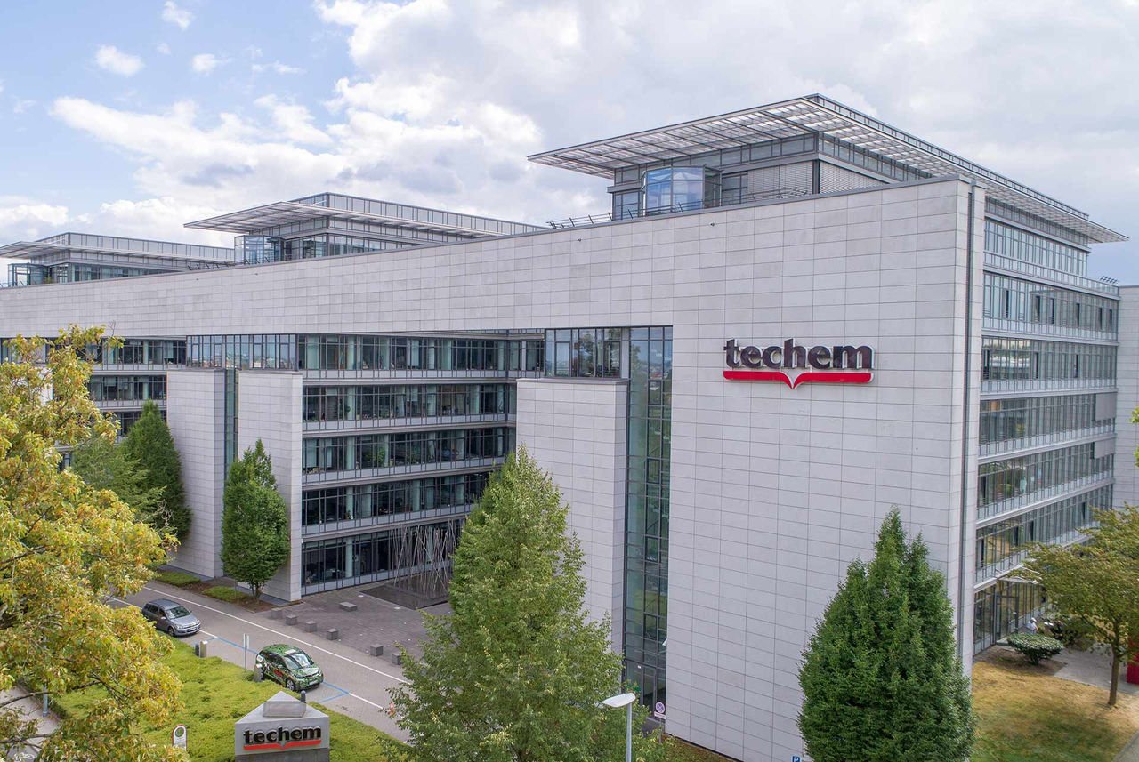 Edificio de oficinas de diseño contemporáneo, identificado claramente por el logotipo rojo y blanco de "techem" ubicado en una de sus fachadas. La estructura presenta líneas arquitectónicas limpias, múltiples plantas con amplios ventanales reflectantes y está construida con paneles de color claro. El edificio se encuentra rodeado de vegetación , incluyendo árboles frondosos, y se aprecian algunos vehículos estacionados en las inmediaciones. El cielo está parcialmente cubierto de nubes, proporcionando una luz diurna suave sobre la escena.