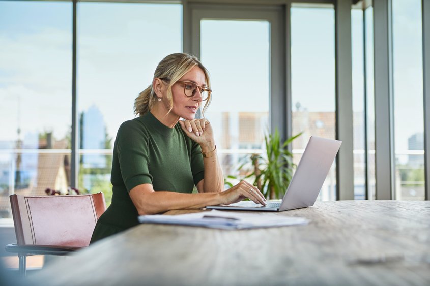 Eine blonde Frau sitzt am Laptop in einem Büro mit großen Glasfronten