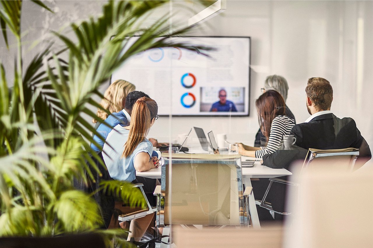 6 Mitarbeitende sitzen in einem verglasten Meetingraum und schauen auf einen Fernseher. Auf dem Fernseher sind verschiedene Grafiken sowie eine weitere Person zu sehen.