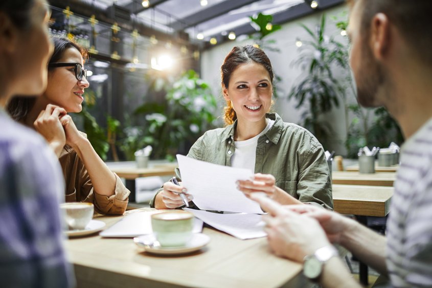 Drei Personen sitzen an einem Tisch in einem Café, eine Person hält Papiere und spricht, auf dem Tisch stehen Tassen und ein Laptop.