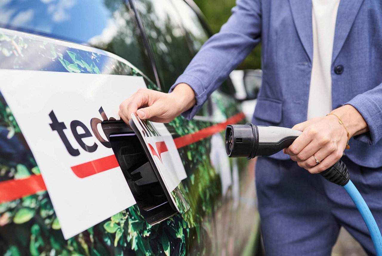 Une femme vêtue d'un costume en lin bleu tient une prise de charge et ouvre la trappe de chargement d'une voiture électrique Techem.