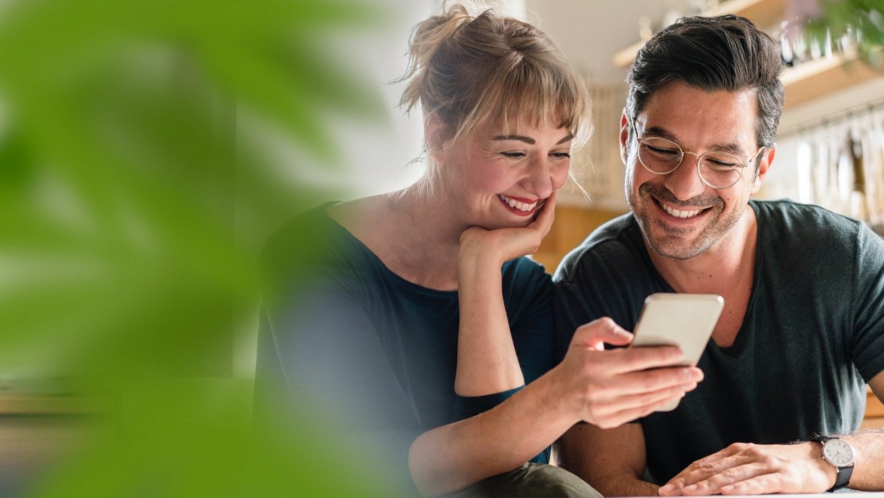 En kvinne som holder en mobiltelefon, mens en mann står ved siden av henne og ser interessert på skjermen. De er omgitt av frodige grønne planter.