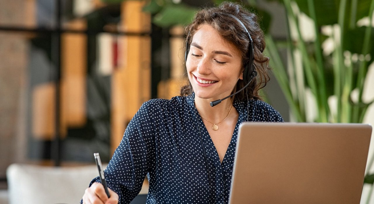 Kvinde sidder med headset og computer og noterer på et stykke papir. 