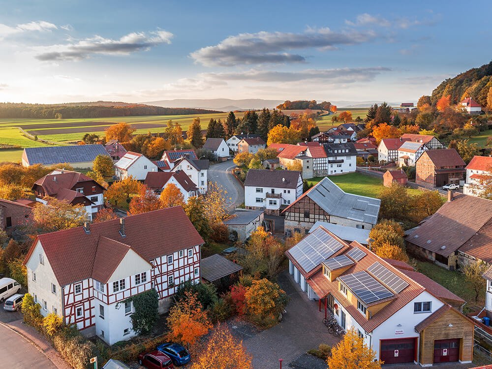 Luftaufnahme eines kleinen Dorfs mit Häusern, teils mit Solaranlagen auf den Dächern. Die Bäume und Felder rundherum zeigen Herbstfarben.