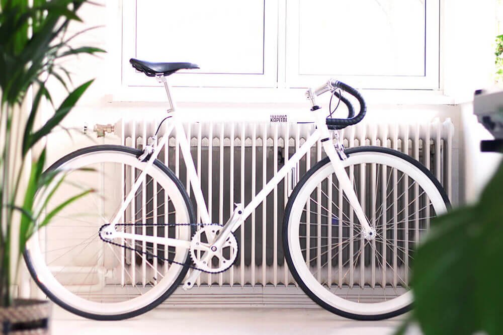 Weißes Fahrrad lehnt an einem Heizkörper unter einem Fenster in einem hellen Raum mit grünen Zimmerpflanzen im Vordergrund.