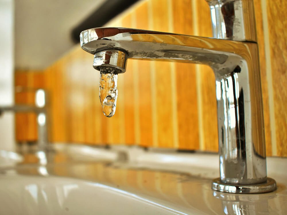 Ein einzelner Wassertropfen hängt an einem metallischen Wasserhahn, kurz bevor er herunterfällt. 