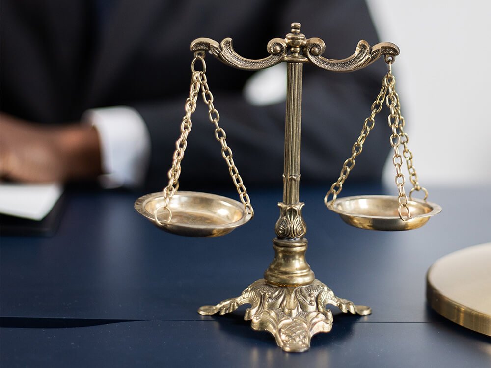 Eine klassische goldene Justitia-Waage steht auf einem dunklen Tisch. Im Hintergrund ist eine Person im Anzug teilweise sichtbar.