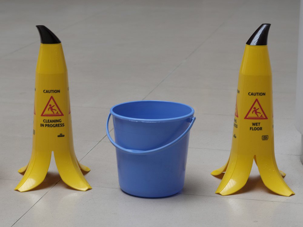 Ein blauer Putzeimer steht auf einem hellen Boden zwischen zwei gelben Warnschildern in Form von Bananenschalen.