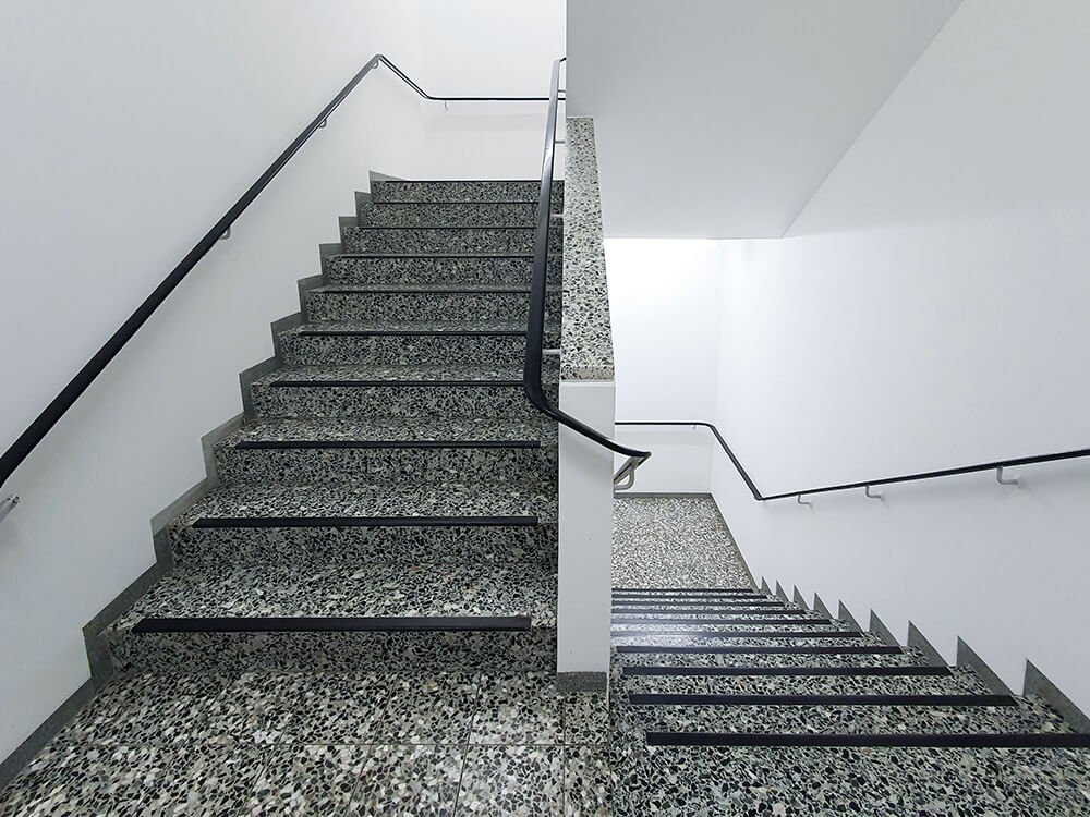 Ein modernes, helles Treppenhaus mit grauen Terrazzo-Stufen, weißen Wänden und schwarzen Handläufen.