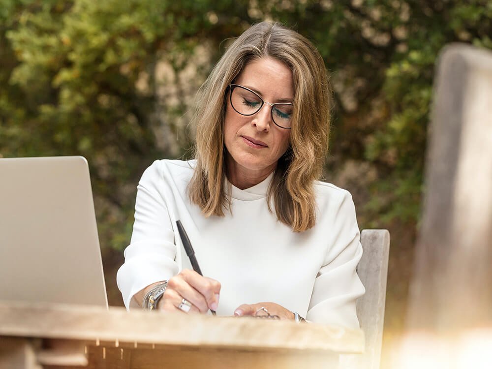 Eine Frau sitzt draußen, schreibt in ein Notizbuch und arbeitet am Laptop.