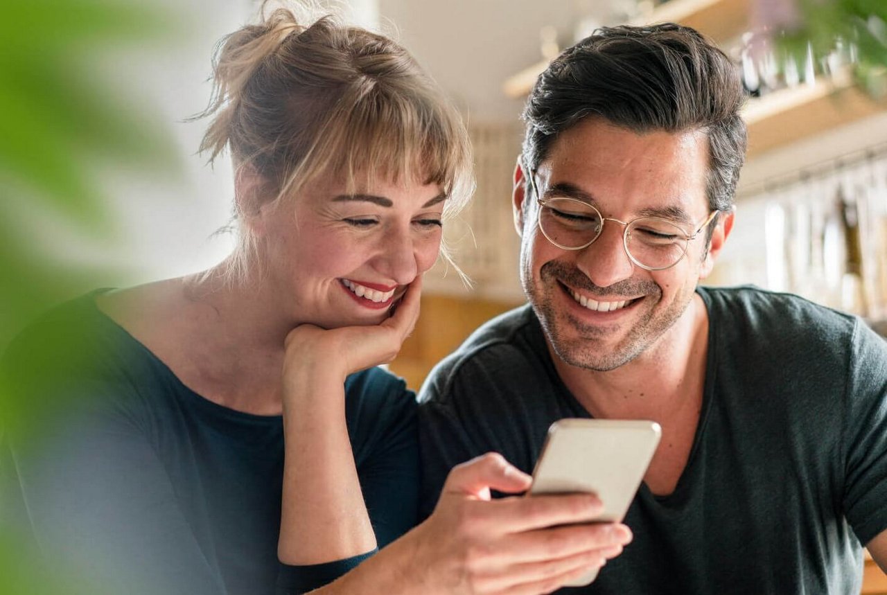 Ein lächelndes Paar sitzt eng beieinander und schaut gemeinsam ein Smartphone. 