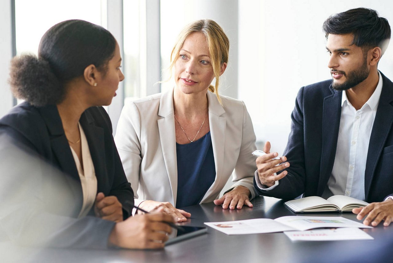 Drei Geschäftspersonen sitzen an einem Tisch in einem modernen Büro. Die Frau in der Mitte spricht, während die anderen zuhören und Notizen vor ihnen liegen.