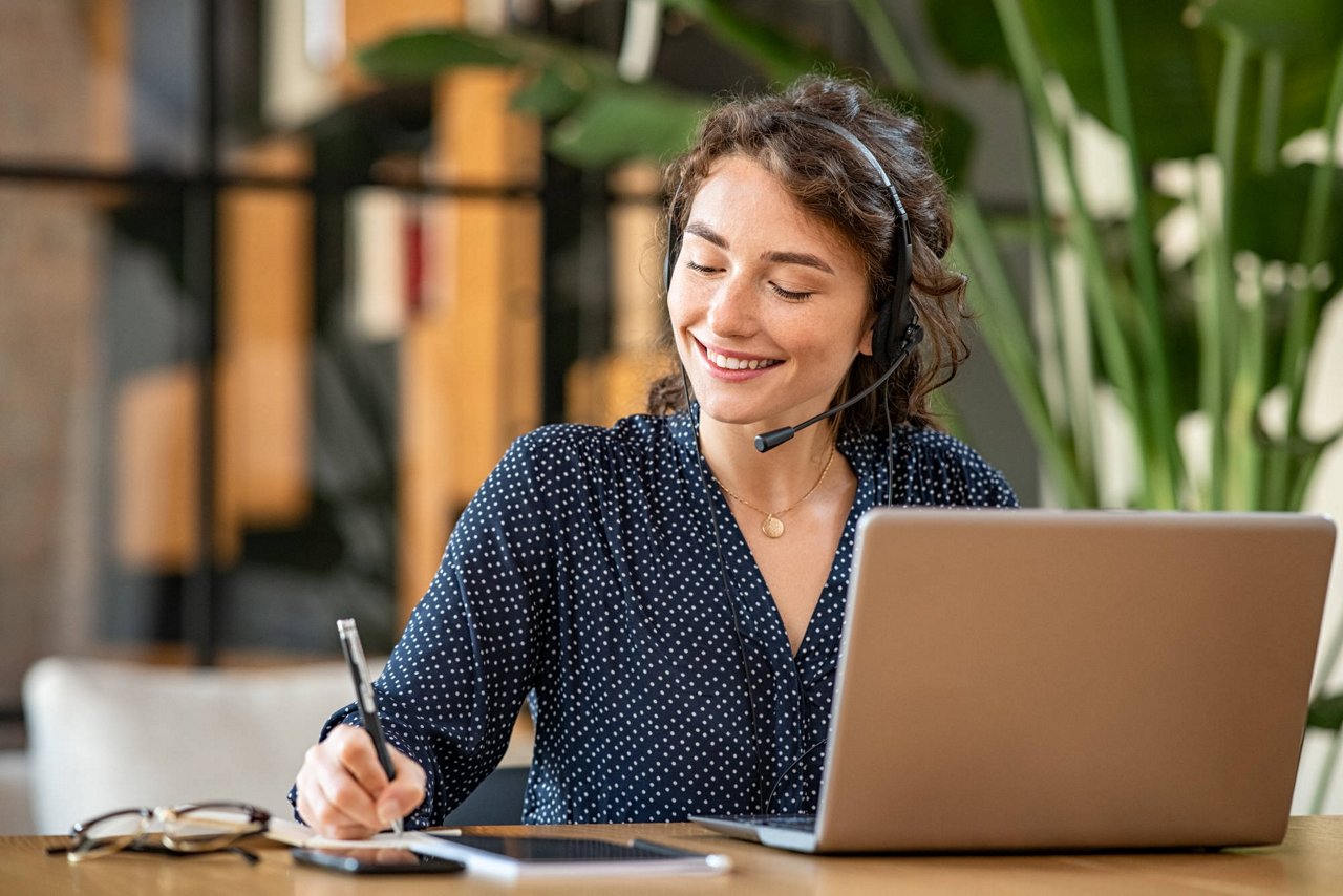 Vendedora de marketing frente a un ordenador, sonriendo mientras atiende una llamada con auriculares y toma notas en un cuaderno