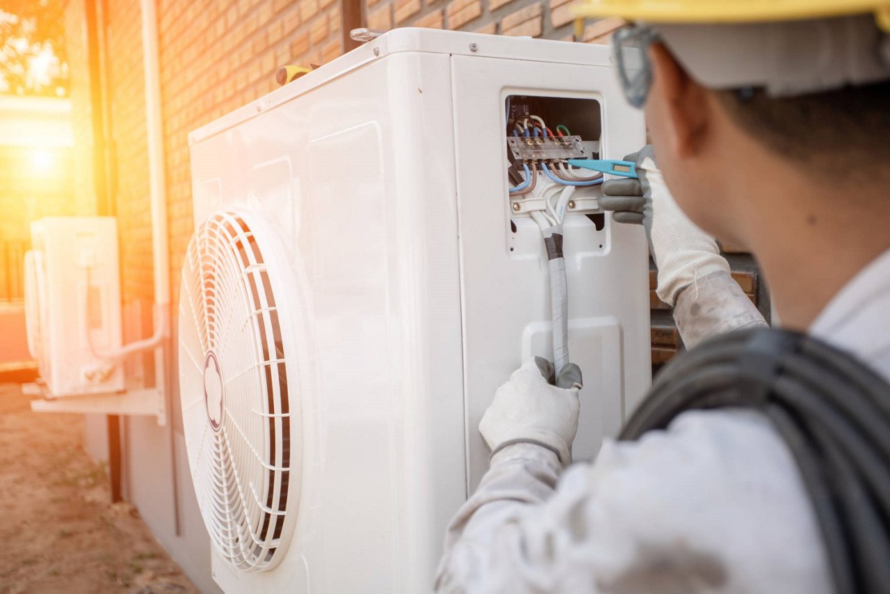 Ein Techniker mit Schutzhelm und Handschuhen arbeitet an der Verkabelung einer Wärmepumpe an einer Hauswand. Die Sonne scheint im Hintergrund, während er mit Werkzeugen die Anschlüsse überprüft.