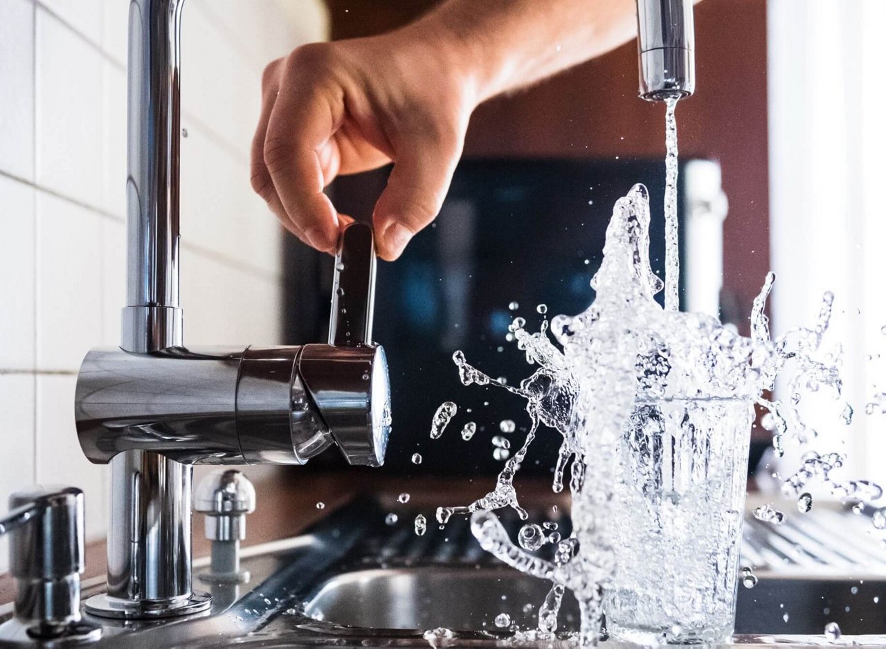 Eine Hand bedient einen Wasserhahn, während Wasser in ein Glas fließt. Das Glas läuft über, und das spritzende Wasser verteilt sich in alle Richtungen über dem Spülbecken. 