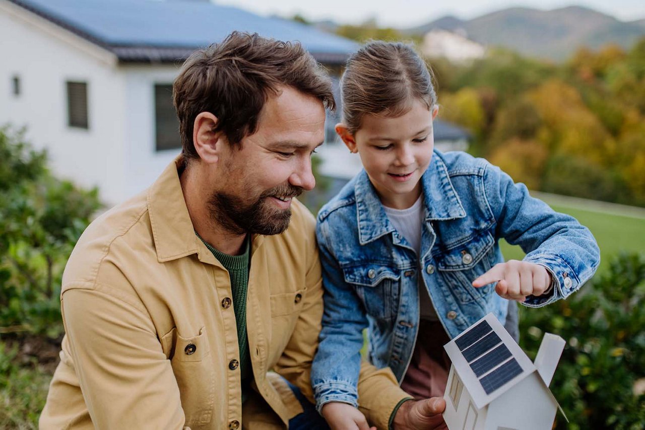 Ein Vater steht mit seiner Tochter lachend im Garten vor einem Haus. Die Tochter hält ein kleines Modellhaus mit Solarpanels in den Händen und zeigt darauf.