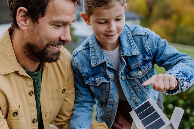 Ein Vater steht mit seiner Tochter lachend im Garten vor einem Haus. Die Tochter hält ein kleines Modellhaus mit Solarpanels in den Händen und zeigt darauf.