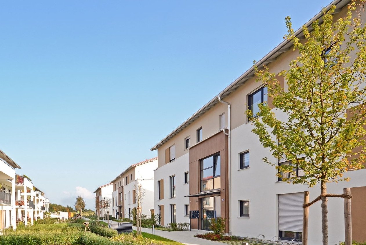 Área residencial moderna con edificios de apartamentos de varios pisos. Los edificios tienen un diseño contemporáneo con grandes ventanas y balcones. Hay un camino bien mantenido y espacios verdes con árboles y arbustos entre los edificios. 