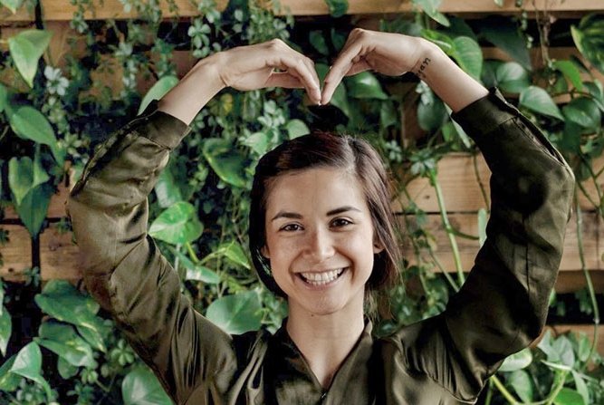  En kvinne som står foran en vegg dekket av planeter, og hun har formet armene sine til et hjerte over hodet. Hun smiler bredt, og uttrykket hennes utstråler glede og positivitet.