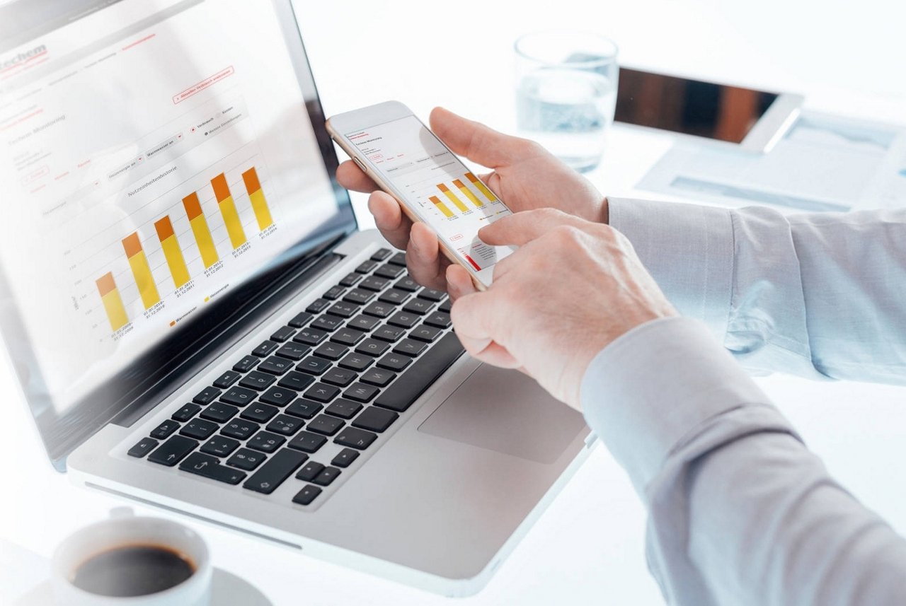 Eine Person analysiert Verbrauchsdaten auf einem Laptop und Smartphone. Die Bildschirme zeigen Balkendiagramme zur Überwachung und Auswertung. Auf dem Schreibtisch liegen eine Kaffeetasse, ein Glas Wasser und ein Tablet.