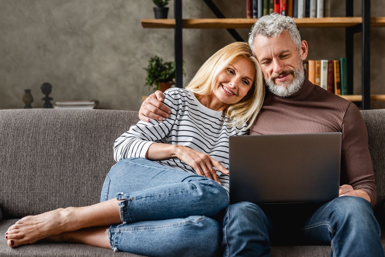 Hombre y mujer abrazados en un sofá, mirando una computadora portátil, con una estantería de libros y adornos en el fondo.