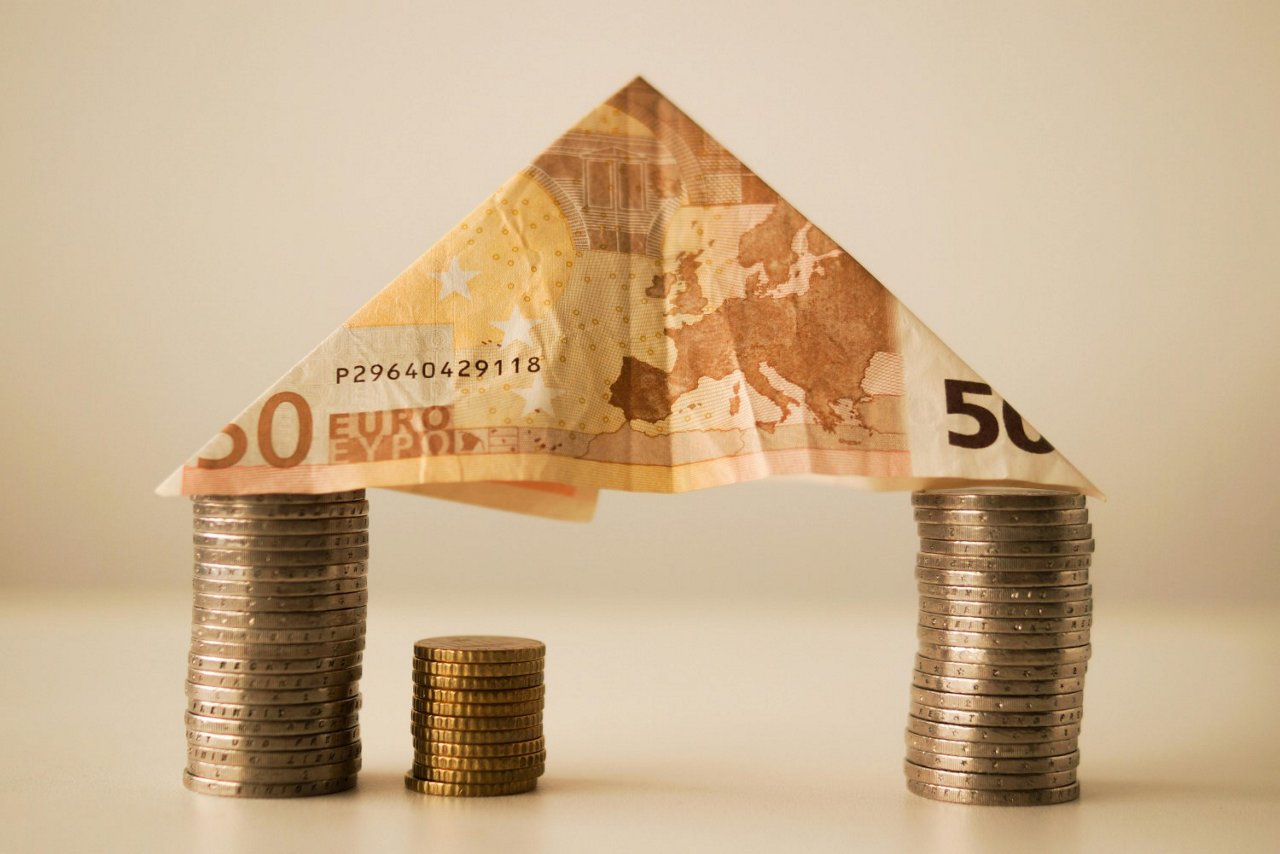 Ein Haus aus Geld: Zwei gestapelte Münztürme als Säulen und ein gefalteter 50-Euro-Schein als Dach, symbolisierend Finanzplanung und Immobilieninvestitionen.