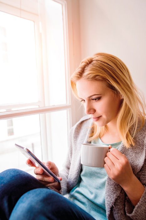 Ragazza bionda seduta vicino alla sua finestra del salotto mentre sta bevendo un caffè e scrollando la home page del suo profilo Instagram.