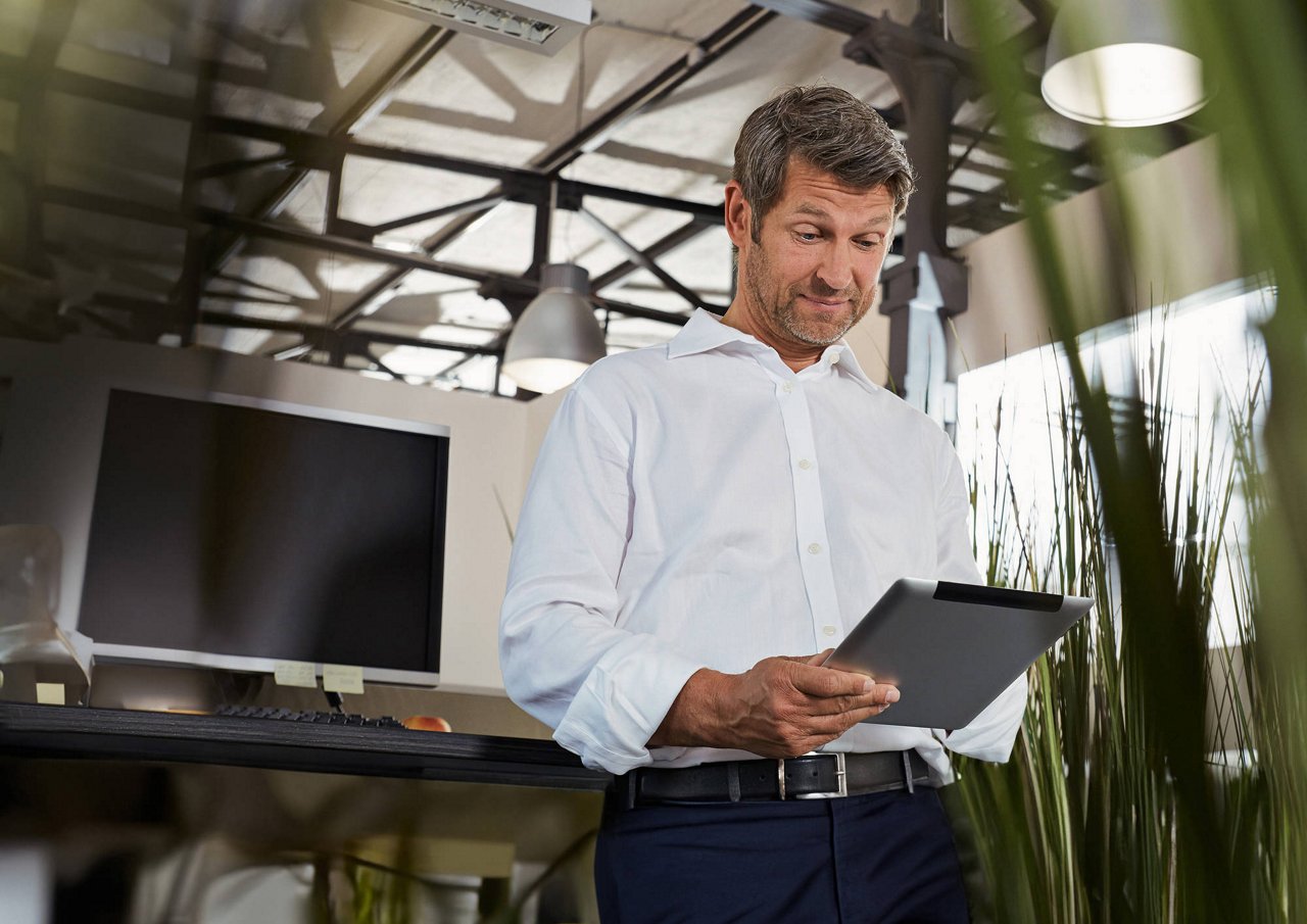 Mann in weißem Hemd liest konzentriert auf einem Tablet in einem modernen Büro mit großem Bildschirm und Pflanzen