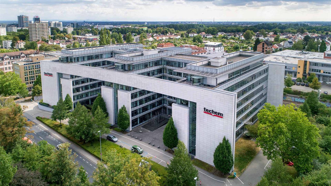 Das Bild zeigt ein Techem Bürogebäude aus Vogelperspektive. Die Fassade ist hell und besteht aus Glas und Stein. Auf dem Gebäude ist das Techem-Logo gut sichtbar. Vor dem Gebäude stehen mehrere Bäume und  Autos.