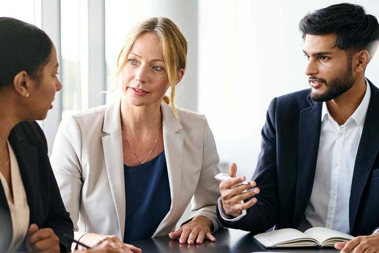 Zwei Frauen und ein Mann in Arbeitskleidung sitzen bei einer Besprechung gemeinsam an einem Tisch. Der Mann hält einen Stift in der Hand und hat ein Notizbuch vor sich liegen.